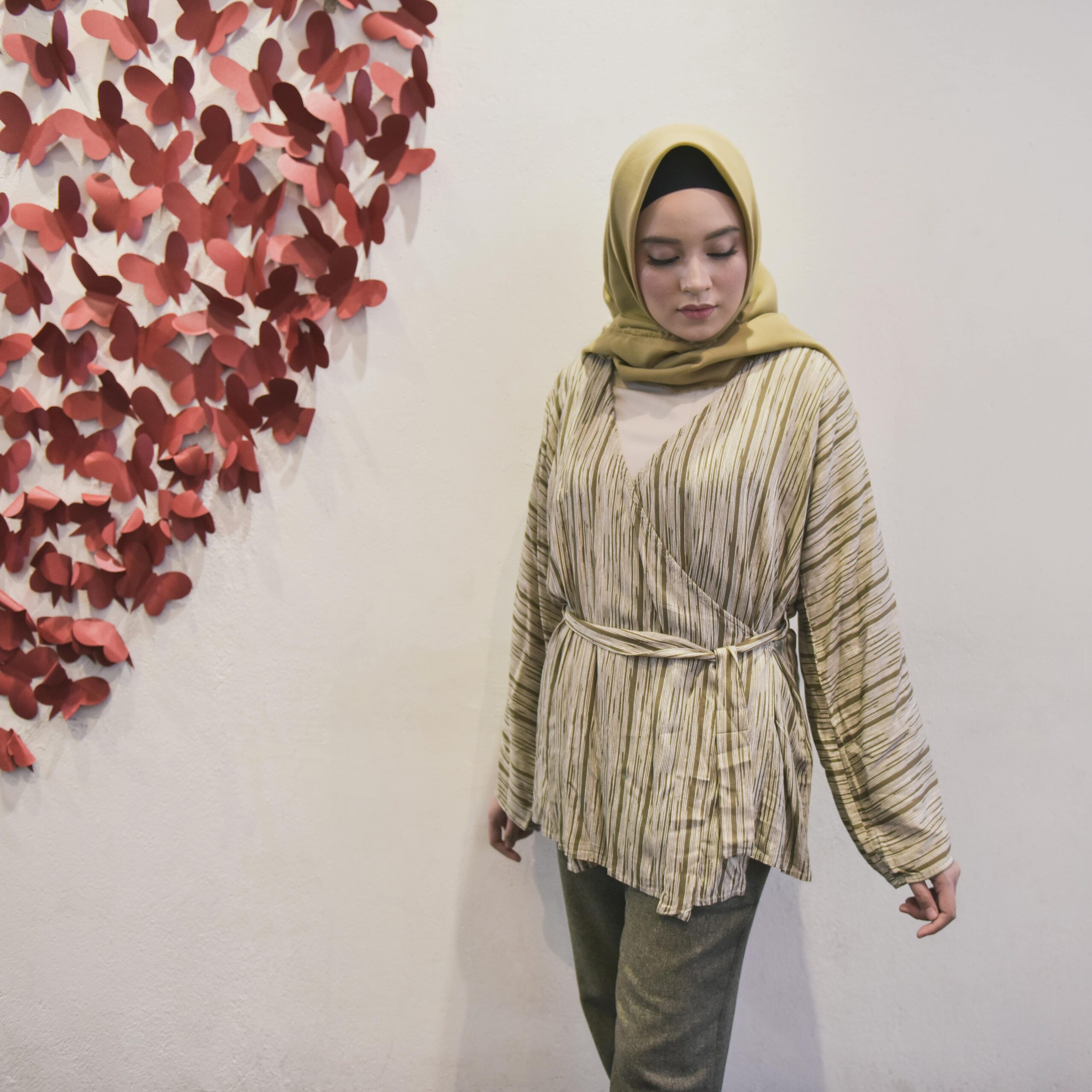 Fashionable woman in hijab standing indoors next to butterfly-shaped wall art installation.